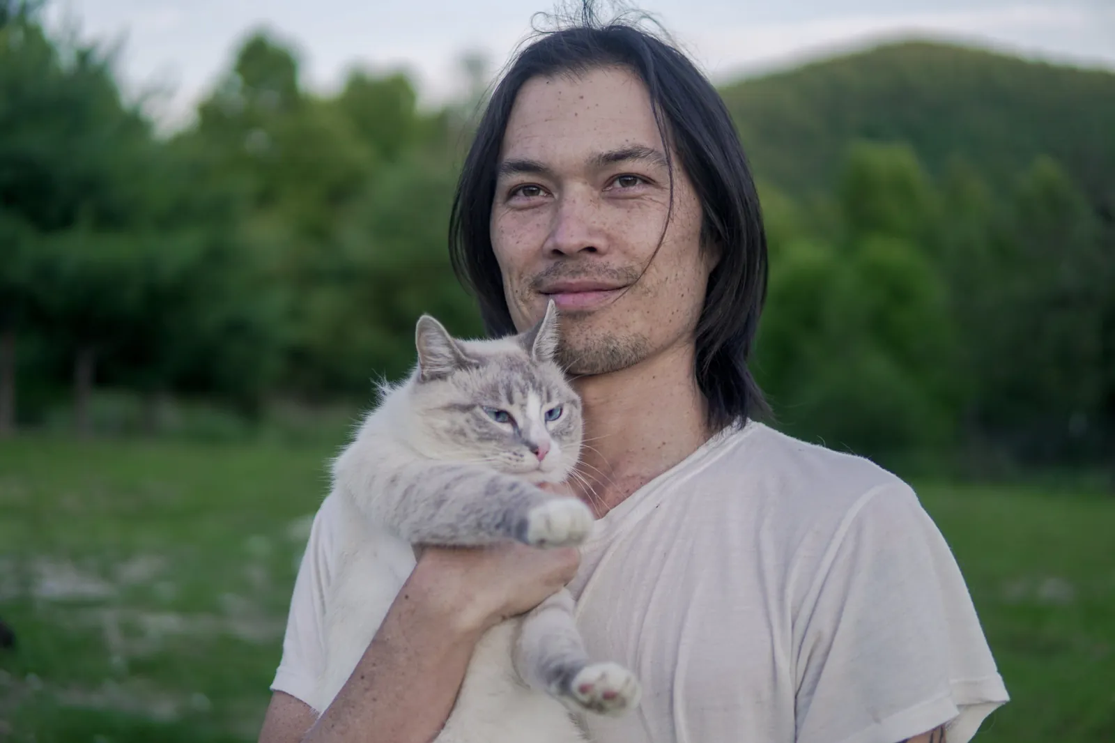Gene Eu holding a grey and white cat in front of a green hillside