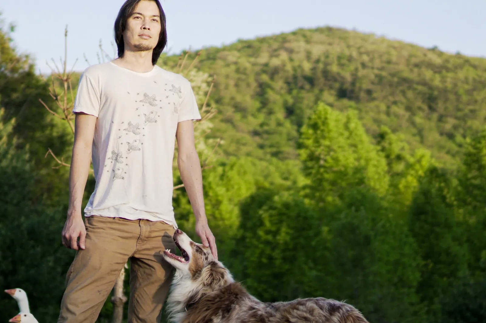 Gene standing in front of a forested mountain with his Australian shepherd and geese